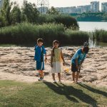 Três crianças brincam perto de um lago em um parque, com edifícios urbanos ao fundo, em um dia ensolarado.