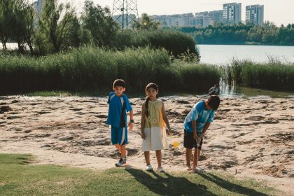 Três crianças brincam perto de um lago em um parque, com edifícios urbanos ao fundo, em um dia ensolarado.