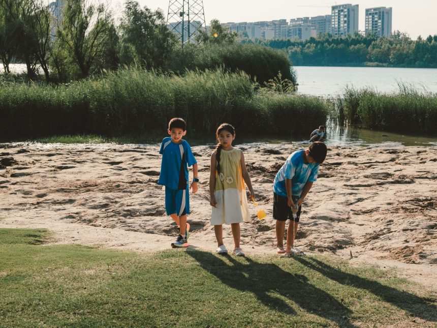 Três crianças brincam perto de um lago em um parque, com edifícios urbanos ao fundo, em um dia ensolarado.