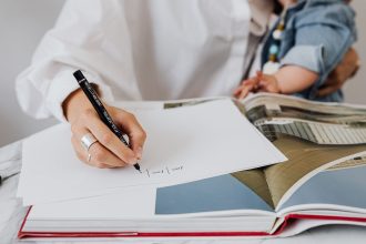 Mãe segura o bebê enquanto escreve em um caderno em ambiente interno.