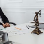 Mesa de trabalho jurídico com uma estátua da Justiça, documentos e um computador portátil.