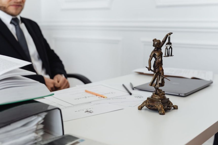 Mesa de trabalho jurídico com uma estátua da Justiça, documentos e um computador portátil.