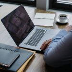 Pessoa usa um notebook sobre uma mesa com documentos e uma xícara de café.