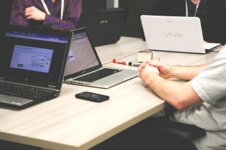 Profissionais participam de uma reunião em uma sala, reunidos ao redor de uma mesa com computadores e cadernos.
